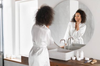 Yüzüne mükemmel bir ten rengi dokunan, günlük cilt bakımı rutinini yapan modern banyo içlerindeki aynanın yanında duran siyahi güzel bir bayan. Güzellik ve şımartma kavramı