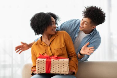 Heyecanlı bir Afrikalı Amerikalı kadın, gülümseyen bir adamdan kalp desenleri olan bir hediye alıyor, güzel aydınlatılmış bir odada neşeli ve samimi bir hediye ve sürpriz anı yaratıyor.