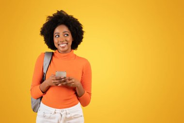 Kıvırcık saçlı, turuncu balıkçı yakalı, akıllı telefonuyla mesajlaşan, gri bir sırt çantası taşıyan, parlak sarı bir arka planı olan mutlu bir genç kadın.