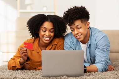 Yerde dizüstü bilgisayarla yatan mutlu Afrikalı Amerikalı çift, kredi kartı tutan kadın, ikisi de rahat ve şık oturma odalarında online alışveriş yapıyorlar.