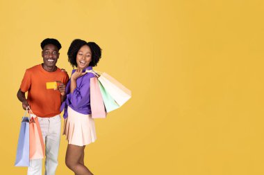 Genç bir çift alışveriş yapıyor, mor bluzlu kadın renkli çantalar tutuyor ve turuncu gömlekli adam kredi kartını gösteriyor, ikisi de sarı arka planda mutlu bir şekilde gülümsüyor.