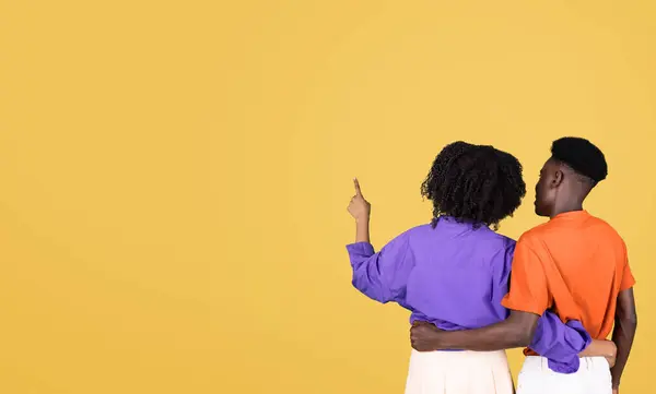 一緒に立っている若いカップルの後ろの景色 オレンジ色のシャツの男性が 平らな黄色の背景に対して 遠くの何かを指している紫色のブラウスの女性 — ストック写真