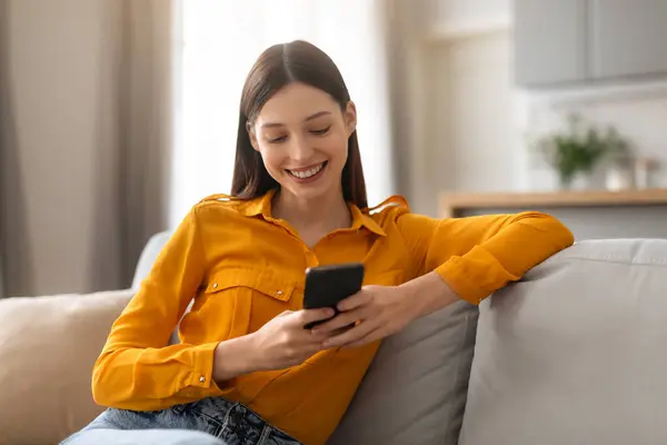 穿着衬衫的松懈的年轻女子喜欢在智能手机上浏览 舒服地躺在灰色沙发上 自然光线照亮着她满意的笑容 — 图库照片