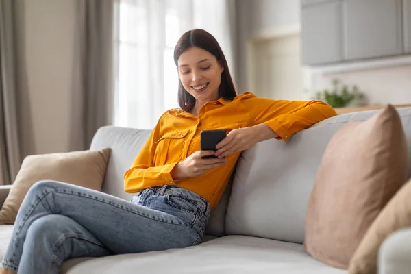 微笑的年轻女子舒服地躺在沙发上 沉浸在智能手机里 身穿鲜艳的黄色衬衫和牛仔裤 坐在舒适 自然光线明亮的客厅里 — 图库照片