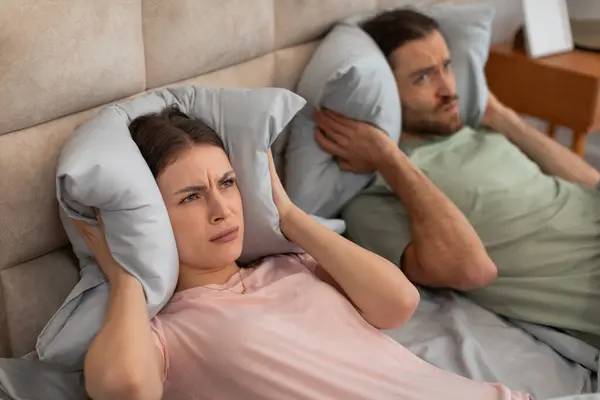 Troubled man and woman in bed cover their ears with soft pillows, visibly upset and seeking relief from an irritating loud noise or snoring, lying in bed
