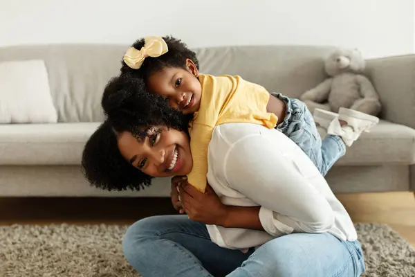 Family portrait of mother and little daughter bonding at home in living room interior. Cute african american toddler child baby girl hugging from behind her beautiful mom, smiling at camera
