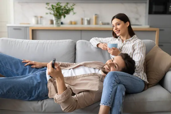 Content man lying on womans lap using remote control and smiling woman holding cup of tea, enjoying cozy moment together on comfortable sofa