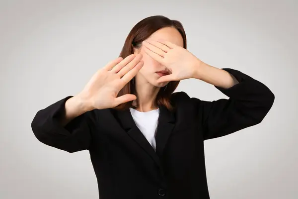 Businesswoman millennial caucasian in a black blazer covers her eyes with her hands, showing a concept of see no evil or avoiding visual input, against a neutral backdrop