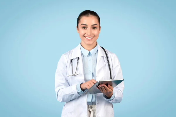 以女医生为重点回顾平板电脑的信息 身穿白衣的女士配戴听诊器 在浅蓝色背景下显示自信 — 图库照片