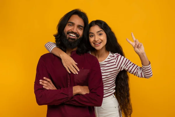 陽気で美しい愛する若いヒンドゥーの夫と妻は黄色いスタジオの背景に結びついています かなりインドの女性は彼女のボーイフレンドを抱きしめ 平和のジェスチャーを示しています — ストック写真
