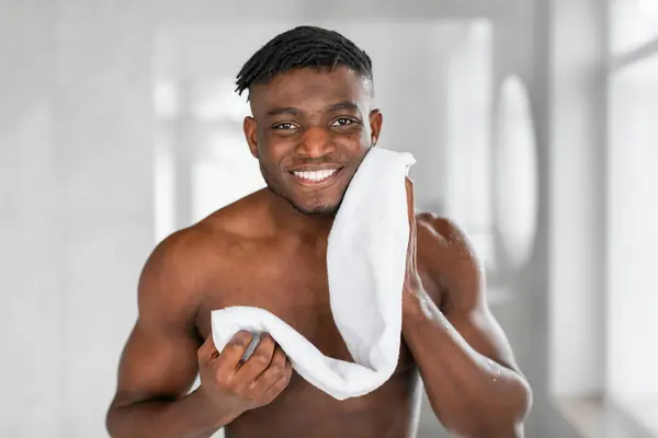 陽気な黒いミレニアル世代の男性の肖像画は 現代のバスルームでタオルで顔を拭き シャツのない立ってカメラに微笑んでいます 男性スキンケアと男性的なグルーミングコンセプト — ストック写真