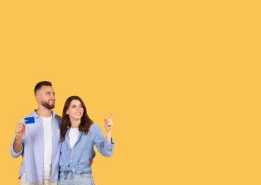Mutlu çift heyecanla yukarı bakıyor, adam kredi kartı sunarken kadın boş yeri işaret ediyor, harcama veya yatırım fırsatları öneriyor.