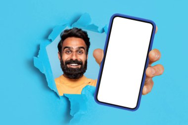 Mutlu bir yetişkin orta doğulu adam cep telefonuyla ekranı boş, mavi stüdyo arka planında bir delik gösteriyor. Uygulama, iş tavsiyesi, kazanma web sitesi