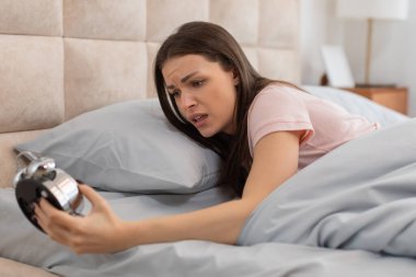 Distressed young woman in bed, frowning at the loud ring of classic alarm clock, capturing the morning struggle of waking up early clipart