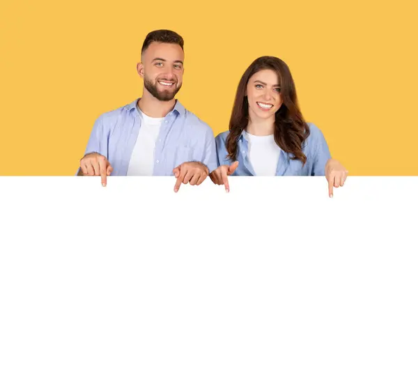 穿着时髦斜纹棉布衬衫的快乐的男女青年指着巨大的空白横幅 横幅上有很大的复制空间 背景是黄色的 — 图库照片