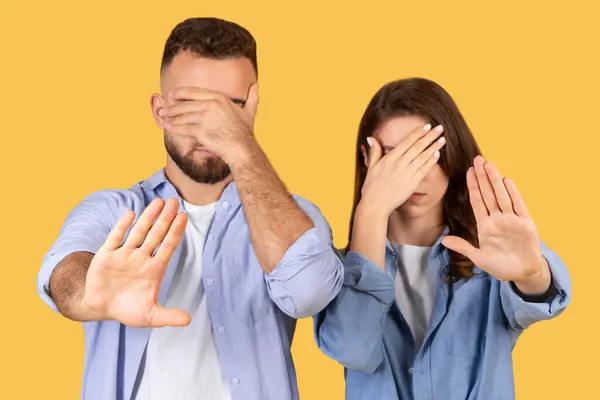 Man and woman covering eyes with one hand while gesturing stop with the other, do not want to see something, standing against yellow background