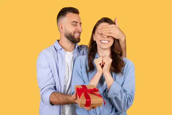 男人用手捂住女人的眼睛微笑 拿着红丝带包裹的礼物 捕捉惊喜和兴奋的瞬间 黄色背景 — 图库照片