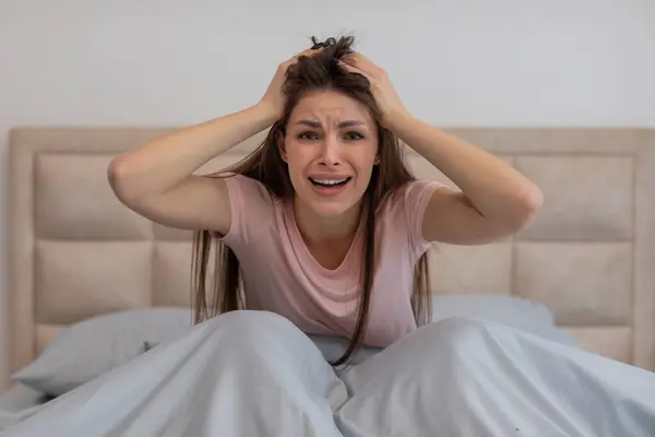 Frustrated young woman in bed gripping her hair with tearful expression of despair, surrounded by crumpled bed sheets in bright room