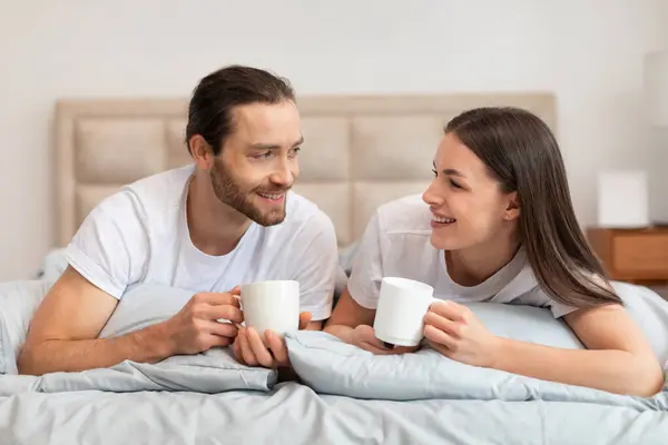 Relaxed and happy couple in white t-shirts enjoying their morning coffee in bed, exchanging smiles and enjoying each others company in serene bedroom