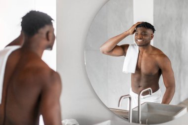 Gülümseyen siyah adam aynaya bakıyor ve duştan sonra saçına dokunuyor, saç bakımına odaklanıyor, modern banyo içinde üstsüz duruyor. Günlük rutin ve kişisel bakım. Seçici odak