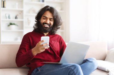 Neşeli, yakışıklı, sakallı, milenyum sakallı Hintli adam evdeki koltukta oturuyor, akıllı telefon ve dizüstü bilgisayar kullanıyor, kopyalama alanı kullanıyor. Gadget bağımlılığı, dijital dünya