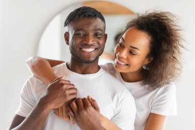 Gülümseyen siyah eşler, modern banyo içinde çekilmiş portre, Afrikalı Amerikalı koca kameraya bakarken karısı onu kucaklıyor, sevginin ve aile içi şımartmanın özünü yakalıyor.