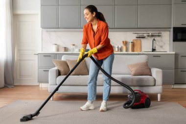 Turuncu gömlekli ve kot pantolonlu neşeli genç bir kadın şık oturma odasında elektrikli süpürge kullanıyor modern gri mobilya ve dekor, tam boy.