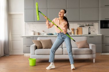 Kulaklıklı öforik kadın neşeli bir şekilde mikrofon olarak yeşil paspası kullanıyor. Parlak ve şık mutfağını temizlerken şarkı söylüyor. Ev işlerini eğlenceyle harmanlıyor.