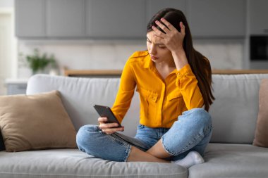 Endişeli genç bir kadın kanepede bağdaş kurmuş oturuyor, akıllı telefonuyla kaşlarını çatıyor, bir eli alnında, yatak haberleri okuyor, modern oturma odasında.