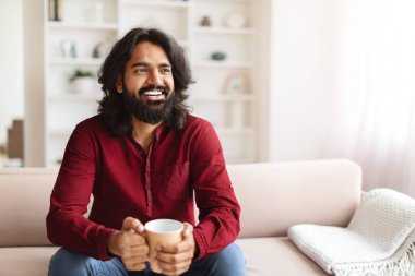 Pozitif yakışıklı sakallı bin yıllık Hintli adam elinde çay fincanıyla kanepede oturmuş reklam için fotokopi odasına bakıyor. Genç doğu çocuğu sabah kahvesinin tadını çıkarıyor.