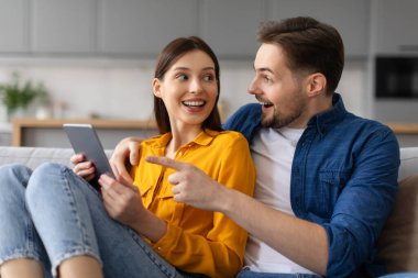 Heyecanlı eşler kanepede oturuyor. Tablet ekranı kadının elinde. Heyecanlı ve keyifli görünüyorlar, internetten alışveriş yapıyorlar.