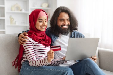 Kolay ödemeler. Bilgisayarlı ve kredi kartlı mutlu Müslüman çift evdeki koltukta dinleniyor, neşeli İslamcı eşler online alışveriş yapıyor, İnternet alışverişlerinin keyfini çıkarıyor,