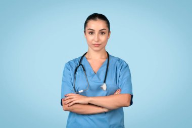 Kolları çaprazlanmış, mavi önlük ve steteskop takan yetenekli genç bayan hemşire mavi arka planda profesyonellik ve hazırlık yayıyor.