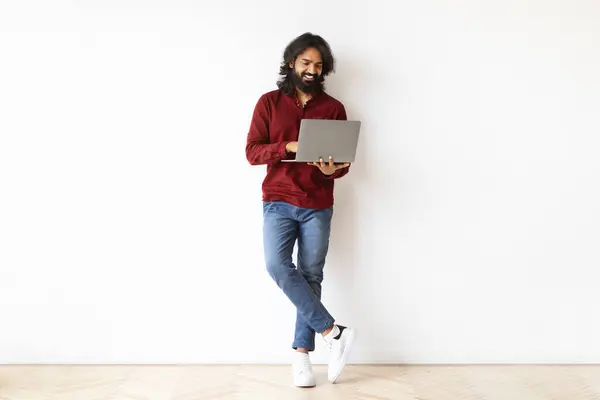 Online learning, e-education for adults. Handsome bearded indian guy using laptop computer on white wall background, checking newest educational course, full length, blank copy space
