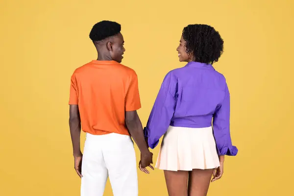 手を握っている若いカップルの背景 オレンジ色のシャツの男と紫色のブラウスの女性は 単純な黄色の背景に対して 肩を振り返ります — ストック写真