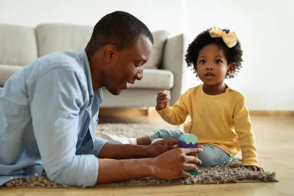 Loving african american dad enjoying development activities with toddler child cute little daughter. Happy black father and kid sitting on floor at home, playing with colorful building blocks