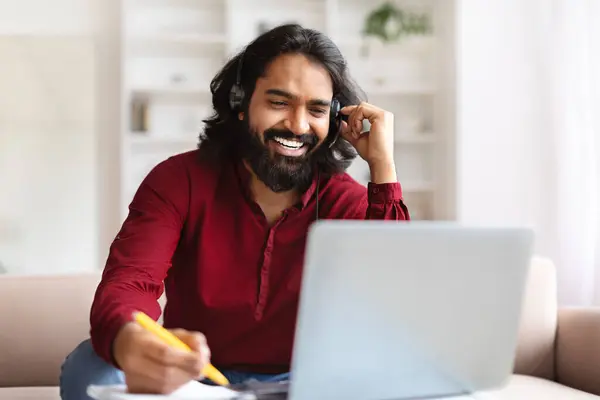 Indian millennial man studying from home, sitting on sofa, looking at laptop screen, using headset, watching online lesson or webinar, taking notes, copy space. Online education for adults