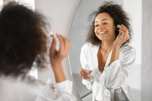 Pretty black woman smiles near mirror while applying facial cosmetic serum, moisturizing skin with natural oils for perfect and healthy complexion, holding dropper near face standing in bathroom