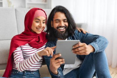 Dijital tabletle gülümseyen Müslüman çift evde yerde dinleniyor, tesettüre bakarken, internette gezinirken veya video izlerken mutlu Arap erkek ve kadın tesettüre girip eğleniyorlar.