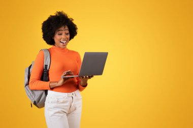 Şaşırtıcı bir şekilde gülümseyen Afro-Amerikan genç kadın heyecanlı bir ifadeyle dizüstü bilgisayara bakıyor, turuncu bir kazak, beyaz bir kot pantolon ve sarı arka planda gri bir sırt çantası var.