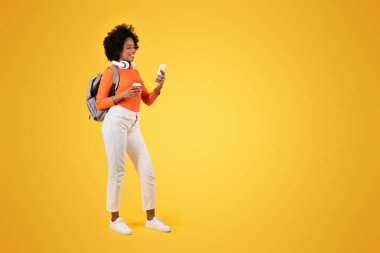 Dalgalı genç bir kadın, akıllı telefonuna bakıyor ve gülümsüyor, beyaz kulaklıklar ve gri bir sırt çantasıyla, turuncu bir bluz ve sarı bir arka planda beyaz pantolon giyiyor.