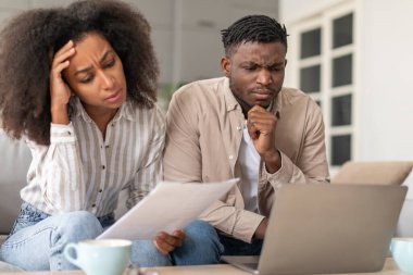 Para sıkıntısı. Kaşlarını çatan Afrikalı evli çift dizüstü bilgisayar kullanıyor, faturalarla ve bütçeyle uğraşıyor, yaşam tarzlarında zorluklarla karşılaşıyor, evdeki mali sorunlarla uğraşıyor.