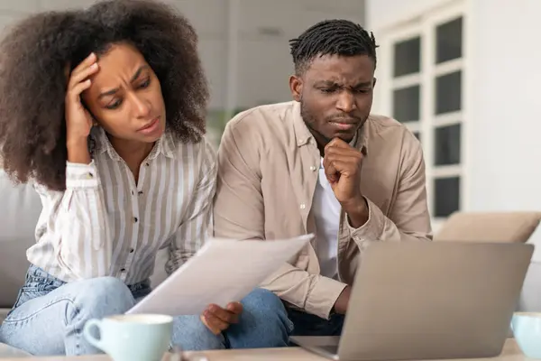 Money troubles. Frowning African American married couple using laptop doing paperwork, working with bills and budgeting, facing challenges in their lifestyle, dealing with financial issues at home