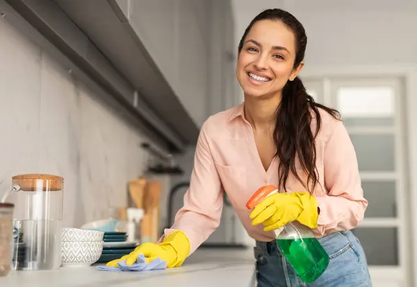 Friendly woman with beaming smile, wearing yellow gloves, cleaning her kitchen counter with blue cloth and green spray bottle, enjoying household routine