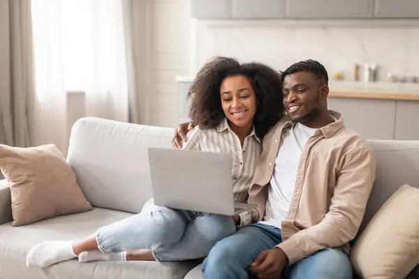 Online movie time. Happy black young spouses sitting with laptop at home, married couple spending quality time together, smiling and watching film on computer seated on sofa indoor