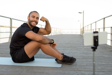 Antrenman sonucu. Spor giyim bölümündeki mutlu sporcu adam, deniz kenarındaki antrenmanını çekerken akıllı telefonuyla pazılarını gösteriyor. Başarılı fitness blog yazma ve refah kavramı