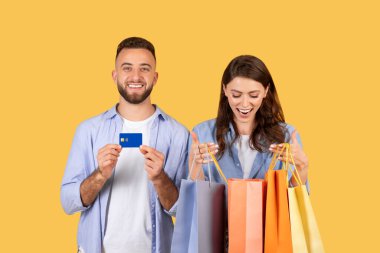 Alışveriş çılgınlığından hoşlanan mutlu genç adam ve kadın, kredi kartı gösteriyor ve birden fazla renkli alışveriş torbası, perakende terapi ve tüketiciliği sembolize ediyor