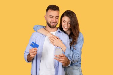 Neşeli çift e-ticaret anlarını paylaşıyor. Elinde kredi kartı olan bir adam ve onu kucaklayan bir kadın. İkisi de akıllı telefona, internetten alışverişe, sarı arka plana bakıyor.
