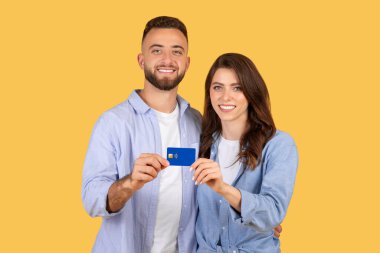 Kredi kartı sunan neşeli genç çift, kameraya özgüvenle bakıyor, finansal güvenlik veya ortak satın alma kararı öneriyor, sarı fon