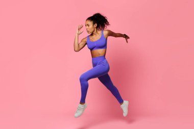 Dinamik fitness. Mor spor giyimli motive olmuş zenci sporcu kadın havada sıçrıyor ve koşuyor. Pembe stüdyo arka planında güç ve atletizm sergiliyor.